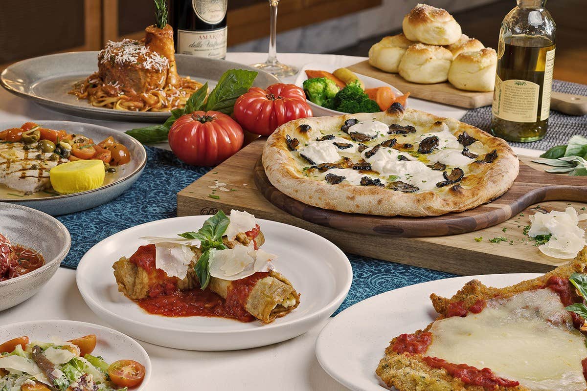 A table filled with various Italian dishes, including pizza, pasta, and appetizers, with wine and fresh vegetables.