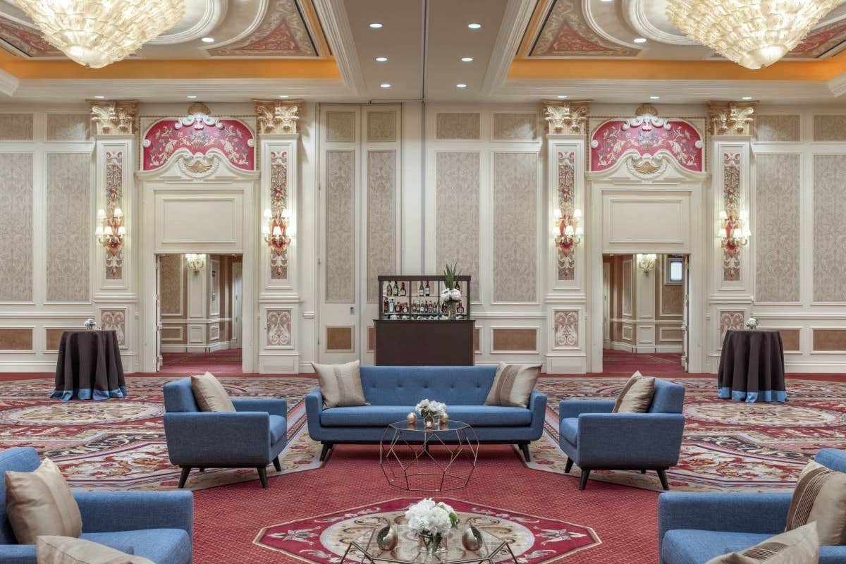 Elegant ballroom with ornate decor, chandeliers, blue sofas, floral arrangements, and a small bar in the background.