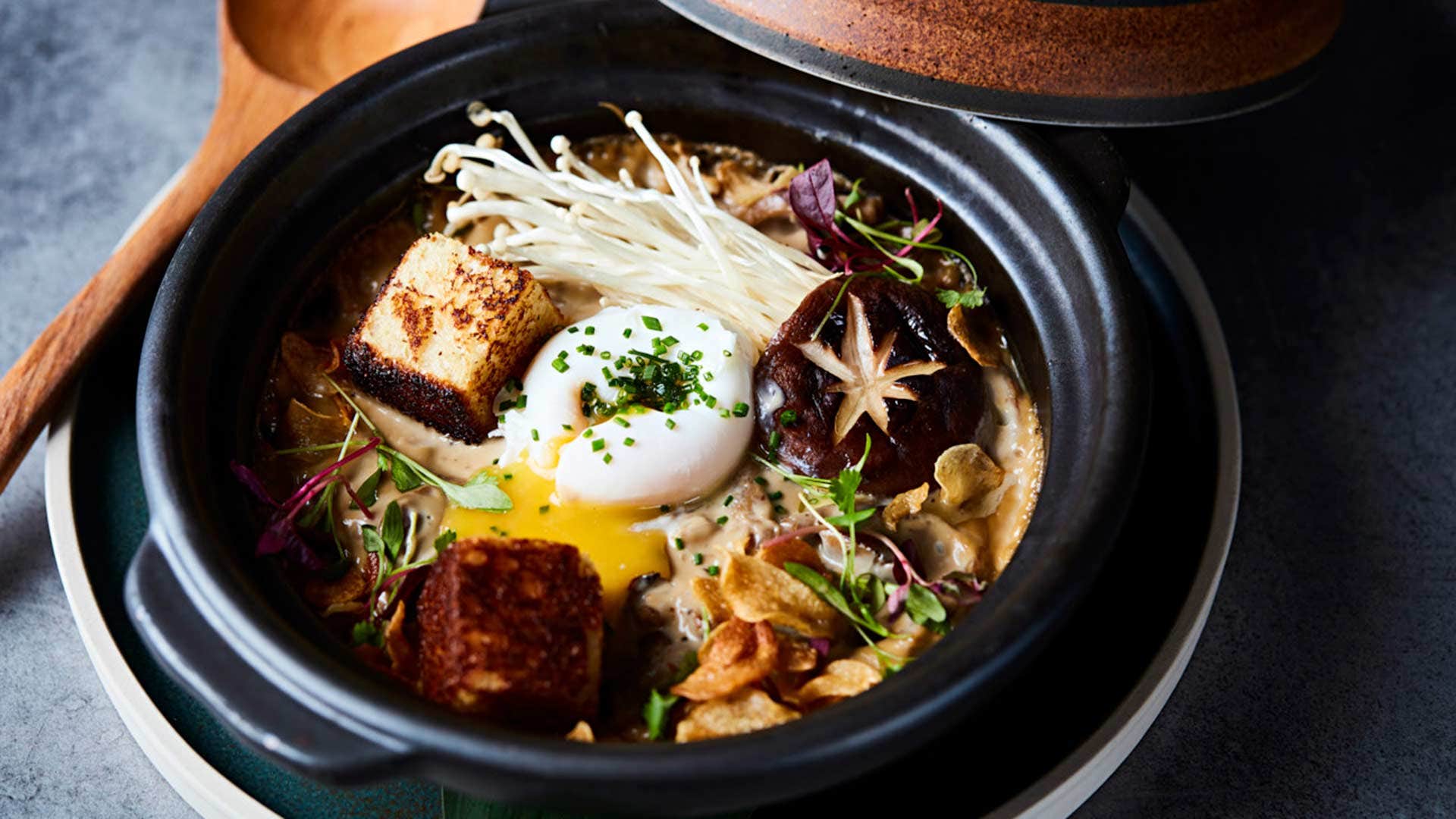 A bowl of gourmet ramen topped with soft-boiled egg, enoki mushrooms, tofu cubes, and various garnishes.